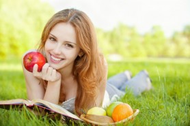 A beautiful young girl with an apple lying on the grass and reading a book beside a basket of fruit is, on a background of green nature